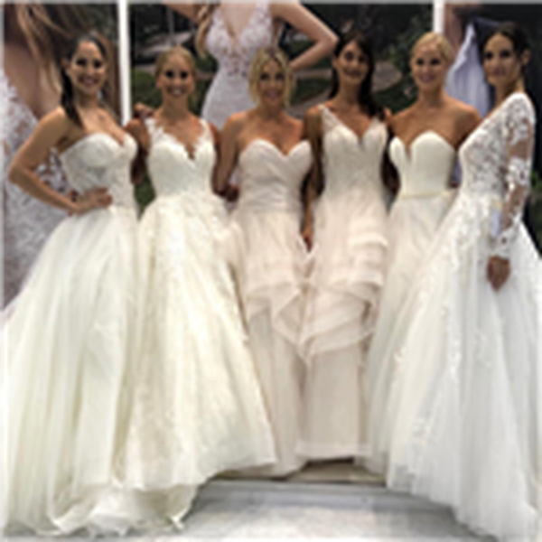 Six female models lined up wearing different wedding dresses from the Ronald Joyce Spring 2020 collection. Models are smiling and linking arms at the launch party. 